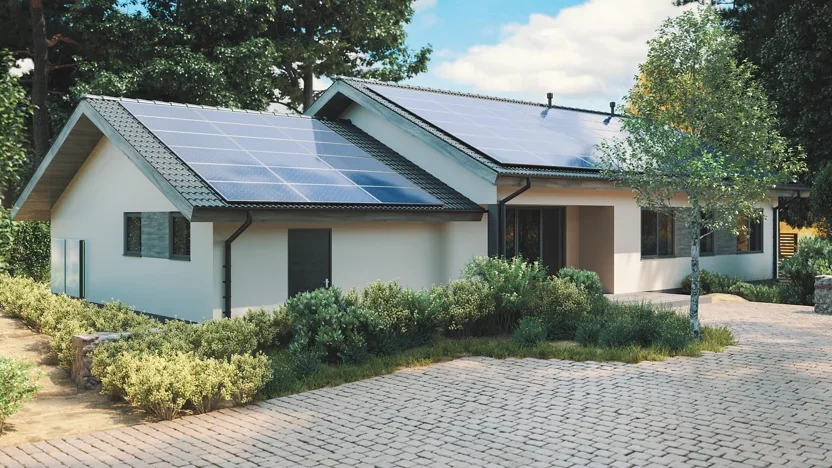 A single-story house with solar panels on its dark gray roof. The house has light-colored walls and is surrounded by lush green bushes and trees. A paved driveway leads to the house.